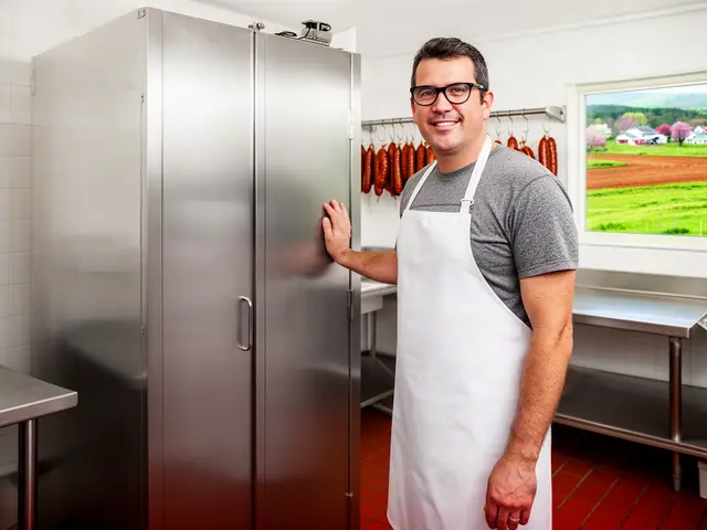 A smiling butcher in a white apron beside a new stainless-steel smoker cabinet with sausages hanging behind.